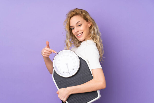Young blonde woman isolated on purple background holding a weighing machine and pointing it