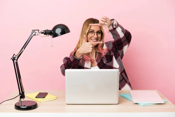 Young student woman in a workplace with a laptop over pink background focusing face. Framing symbol