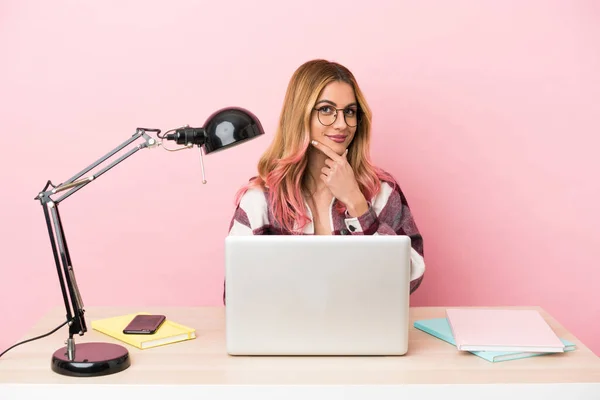 Young student woman in a workplace with a laptop over pink background thinking