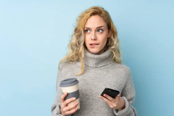 年轻的金发女人穿着一件蓝色背景的毛衣 一边拿着咖啡 一边想着什么 一边拿着手机 — 图库照片