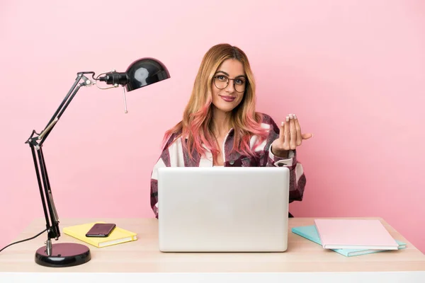 Young student woman in a workplace with a laptop over pink background inviting to come with hand. Happy that you came