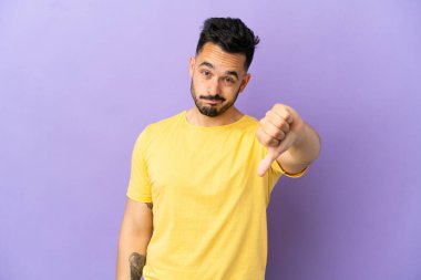 Mor arka planda izole genç beyaz adam olumsuz ifade ile başparmağını aşağı gösteriyor
