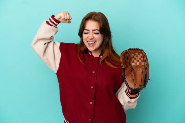 Beyzbol oynayan genç kızıl saçlı kadın mavi arka planda izole edilmiş güçlü bir jest yapıyor.