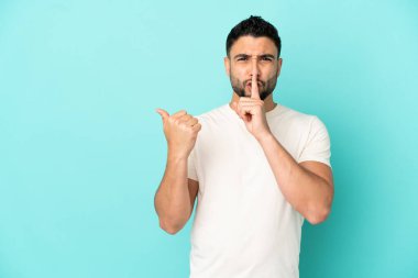 Genç Arap adam mavi arka planda izole edilmiş bir şekilde kenarı işaret ediyor ve sessizlik jesti yapıyor.