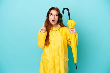 Kızıl saçlı kadın elinde şemsiyeyle mavi arka planda izole edilmiş bir şekilde parmağını kaldıran bir fikir düşünüyor.