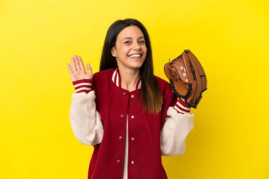 Beyzbol oynayan beyaz bir kadın. Sarı arka planda izole edilmiş. Mutlu bir ifadeyle selam veriyor.
