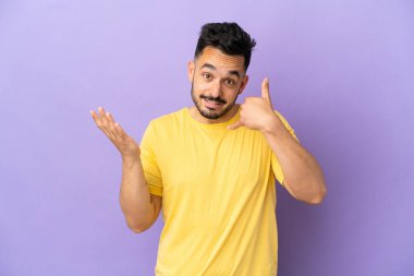 Mor arka planda izole edilmiş genç beyaz bir adam telefon jesti yapıyor ve şüpheli.