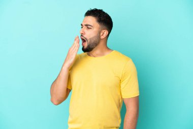 Genç Arap adam mavi arka planda izole edilmiş esniyor ve ağzını eliyle açık tutuyor.