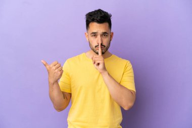 Mor arka planda izole edilmiş genç beyaz adam yan tarafı işaret ediyor ve sessizlik hareketi yapıyor.