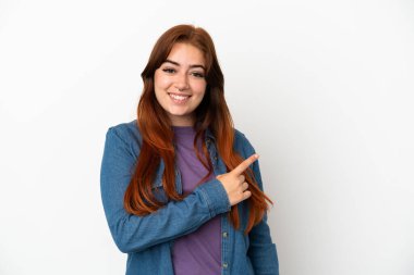 Beyaz arka planda izole edilmiş genç kızıl saçlı kadın bir ürünü sunmak için kenarı işaret ediyor.