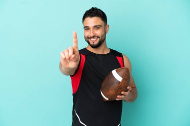 Genç Arap rugby oynayan adam mavi arka planda izole edilmiş ve parmağını kaldırıyor.