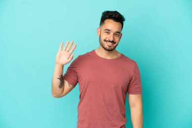Mavi arka planda izole edilmiş genç beyaz adam mutlu bir ifadeyle selamlıyor.