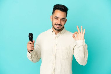 Beyaz tenli genç şarkıcı, mavi arka planda izole edilmiş. Parmaklarıyla tamam işareti gösteriyor.