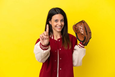 Beyzbol oynayan beyaz bir kadın sarı arka planda izole edilmiş ve parmağını kaldırıyor.