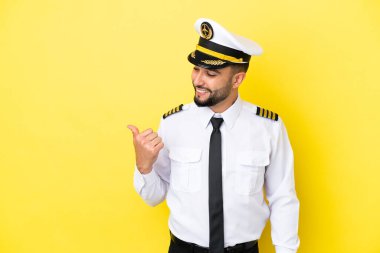 Arap uçak pilotu, sarı arka planda izole edilmiş bir şekilde bir ürün sunmak için kenarı işaret ediyor.