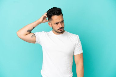 Kafkasyalı genç adam mavi arka planda izole edilmiş. Kafası kaşınırken şüpheleri var.
