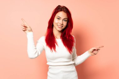 Pembe arka planda izole edilmiş genç kızıl saçlı kız parmak uçlarını gösteriyor ve mutlu.