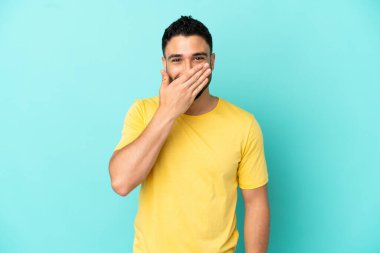 Genç Arap adam mavi arka planda izole edilmiş mutlu ve gülümseyen ağızlarını eliyle kapatıyor.