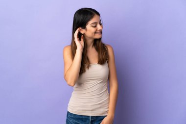 Mor arka planda izole edilmiş genç beyaz kadın elini kulağına koyarak bir şeyler dinliyor.