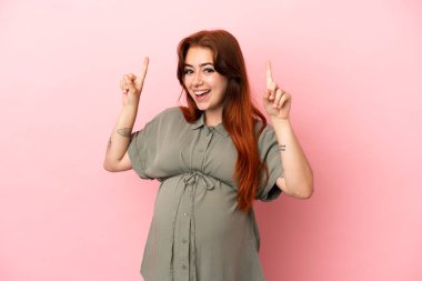 Genç kızıl saçlı beyaz kadın pembe arka planda izole edilmiş hamile ve yukarı bakıyor.
