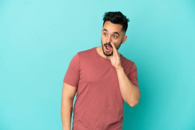 Mavi arka planda izole edilmiş genç beyaz adam yan tarafa bakarken sürpriz bir hareketle bir şeyler fısıldıyor.