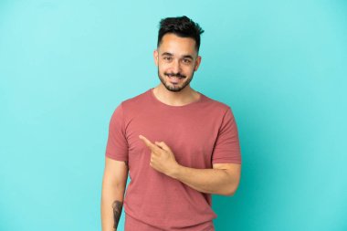 Mavi arka planda izole edilmiş genç beyaz adam bir ürünü sunmak için kenarı işaret ediyor.