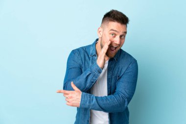 Mavi arka planda izole edilmiş Rus yakışıklı bir adam bir ürün sunmak ve bir şeyler fısıldamak için kenarı işaret ediyor.