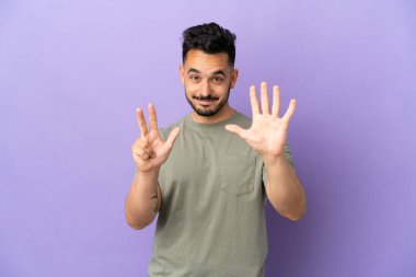 Mor arka planda izole edilmiş genç beyaz adam parmaklarıyla sekiz sayıyor.