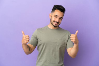 Mor arka planda izole edilmiş genç beyaz bir adam baş parmak hareketi yapıyor.