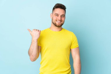 Mavi arka planda izole edilmiş Rus yakışıklı bir adam bir ürün sunmak için kenarı işaret ediyor.