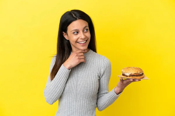 Young Caucasian Woman Holding Burger Isolated Yellow Background Looking While — Stock Photo, Image