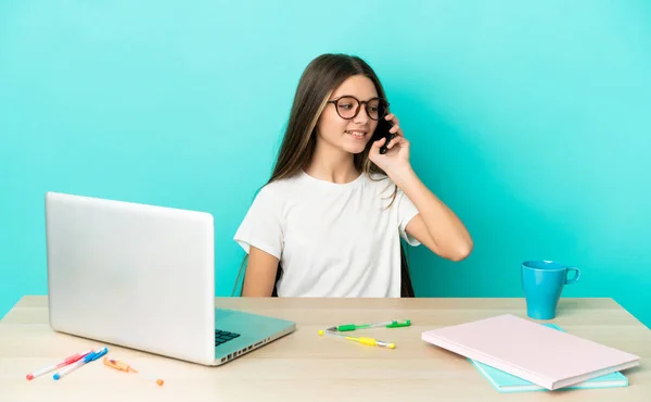 Little girl in a table with a laptop over isolated blue background keeping a conversation with the mobile phone