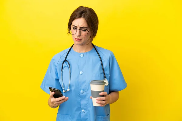 Surgeon doctor woman isolated on yellow background holding coffee to take away and a mobile