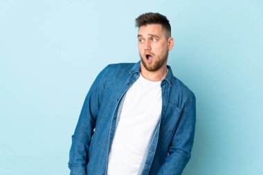 Mavi arka planda izole edilmiş Rus yakışıklı adam yan tarafa bakarken sürpriz jest yapıyor.
