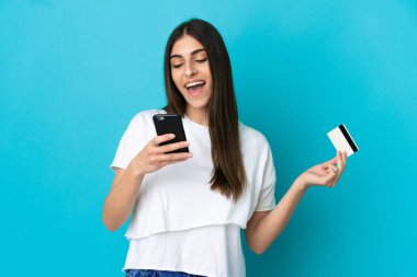 Genç beyaz kadın mavi arka planda izole edilmiş cep telefonuyla kredi kartıyla alışveriş yapıyor.