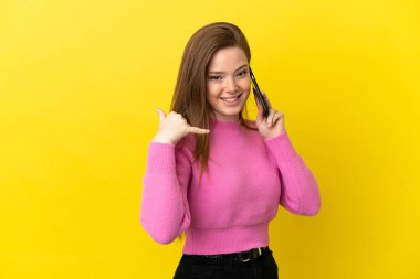 İzole edilmiş sarı arka planda cep telefonu kullanan genç bir kız telefon hareketi yapıyor. Bana geri dönüş işareti ver.