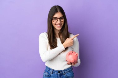 Mor arka planda izole edilmiş domuz kumbarası tutan beyaz bir kadın bir ürünü sunmak için yan tarafı işaret ediyor.