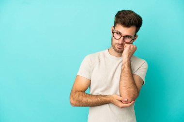 Mavi arka planda soyutlanmış, yorgun ve sıkılmış bir ifadeyle genç, beyaz, yakışıklı bir adam.