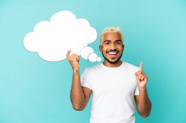 Genç Kolombiyalı yakışıklı adam mavi arka planda izole edilmiş ve düşünce balonu tutuyor.