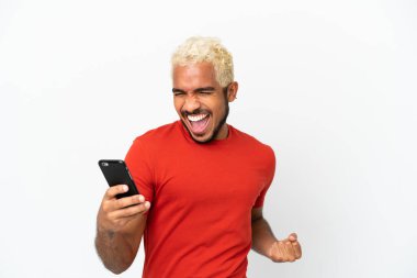 Genç Kolombiyalı yakışıklı beyaz arka planda izole edilmiş cep telefonu kullanıyor ve zafer jesti yapıyor.