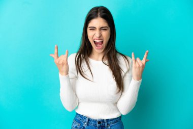 Mavi arka planda izole edilmiş genç beyaz kadın boynuz hareketi yapıyor.