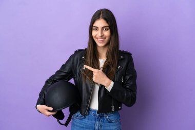 Mor arka planda izole edilmiş motosiklet kasklı beyaz bir kadın bir ürünü sunmak için yan tarafı işaret ediyor.