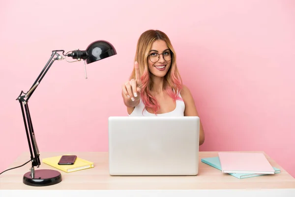Young student woman in a workplace with a laptop over pink background showing and lifting a finger