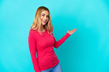 İzole edilmiş mavi arka planda genç bir kadın. Davet etmek için ellerini yana uzatıyor.