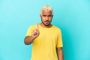 Mavi arka planda izole edilmiş genç Kolombiyalı yakışıklı adam ciddi bir ifadeyle birini sayıyor.