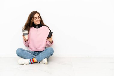 Beyaz arka planda izole bir şekilde yerde oturan beyaz tenli genç bir kadın kahve alıp götürmek için elinde bir cep telefonu...