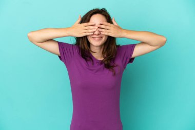 Mavi arka planda izole edilmiş genç beyaz bir kadın. Gözleri elleriyle kaplıyor ve gülümsüyor.
