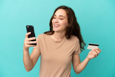 Genç beyaz kadın mavi arka planda izole edilmiş cep telefonuyla kredi kartıyla alışveriş yapıyor.
