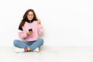 Beyaz arka planda zemin üzerinde oturan beyaz tenli genç kadın telefon zafer pozisyonunda.