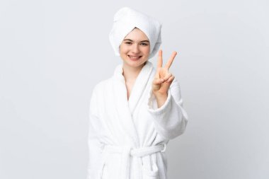 Beyaz arka planda izole edilmiş bornozlu genç kız gülümsüyor ve zafer işareti gösteriyor.